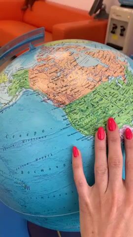The video shows a close-up view of a globe with a woman's hand with bright pink nail polish gently touching and rotating it. The globe displays continents and oceans, with visible country borders and names, some of which appear to be in Cyrillic script, indicating it is likely a Russian-language globe. The woman's hand is positioned over North America initially, then moves to show parts of the Atlantic Ocean and Europe/Middle East. The background is blurred but suggests an indoor setting with an orange sofa and office equipment. The lighting is bright, and the focus remains primarily on the globe and the woman's hand.