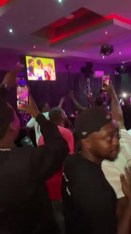 The video shows a group of people gathered inside a club or bar. A TV screen shows a sporting event, possibly a soccer match, with players in the background. The people are watching the screen and cheering, raising their arms in excitement, and filming with their phones. The lighting in the club is dim, with colorful LED lights illuminating the ceiling, walls, and the bar area. The camera moves around, capturing the crowd's reaction to the game. The atmosphere is lively and celebratory.