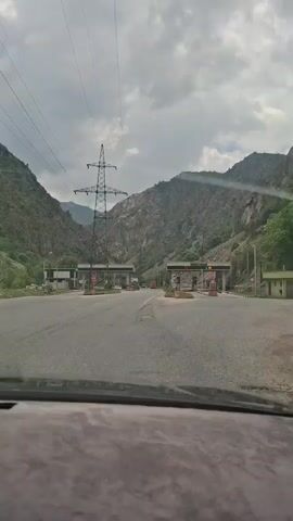 The video shows a view from inside a car driving on a road with toll booths. The road is flanked by mountains. A tall power line tower is visible on the left side of the road. The sky is cloudy. There is a light amount of traffic at the toll booths.