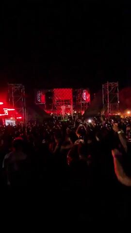A nighttime outdoor concert is depicted. A large crowd of people are facing a stage with a complex lighting and sound system. Bright red and white lights illuminate the stage and the smoke effects. Many people in the crowd are holding up their mobile phones, likely recording the performance. The overall atmosphere appears to be energetic and celebratory.