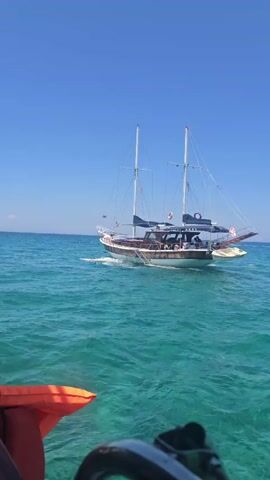A boat with two masts sails on turquoise water under a clear blue sky. The boat has a wooden hull with several people visible on its deck. A bright orange object, possibly a kayak, is in the foreground, partially obscuring the view of the boat.
