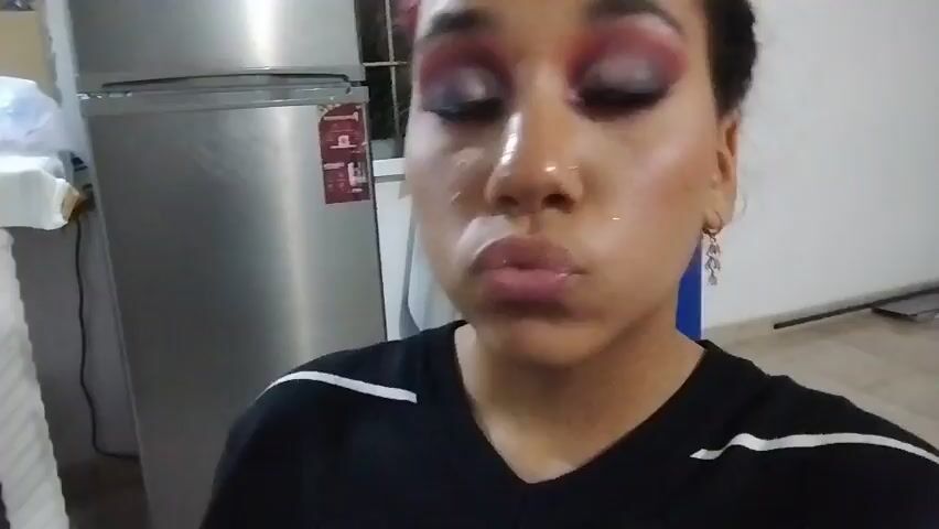 A medium shot shows a woman with dark hair and heavy eye makeup. Her left hand is pressed against her cheek, and she is sticking out her tongue. She then makes a pouting face. The background includes a stainless steel refrigerator and a tiled floor.
