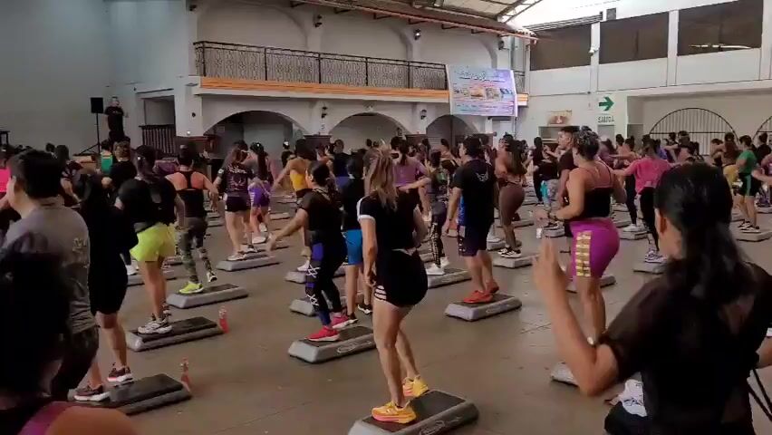 A large group of people, predominantly women, are participating in a fitness class, likely aerobics or step aerobics. They are arranged in rows on a spacious, indoor floor, each person standing on an individual grey aerobic step. The individuals are dressed in athletic wear, including leggings, shorts, and sports bras. The lighting appears to be artificial, and the environment resembles a gymnasium or a large hall, with a high ceiling and balconies visible in the background. A man is standing on a platform to the left of the frame, possibly leading the class. A sign with text and images is displayed on a wall in the background, and an exit sign with a green arrow is also visible. Some individuals are captured mid-movement, suggesting the video is documenting an active exercise session. Water bottles and gym bags are scattered on the floor. The overall scene depicts a communal fitness activity in an open indoor space.