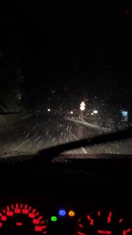 The video depicts a night-time driving scene during heavy snowfall. The view is from inside the car, looking out through the windshield. The road ahead is visible, illuminated by the car's headlights and streetlights.  Snow is falling heavily and being cleared by the windshield wipers. Speed limit signs are visible on the side of the road. The car's dashboard is visible, showing the speedometer, RPM gauge, and indicator lights.  The environment appears to be a mountainous or hilly area with snow-covered trees.