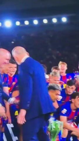 A group of soccer players wearing matching blue and red jerseys are gathered together, cheering and lifting a large silver trophy. The trophy is adorned with red and blue ribbons, and the players are raising their arms in celebration. They are surrounded by a crowd of people in the background, and bright lights are illuminating the scene. One man in a blue suit can be seen walking off camera to the left. The overall atmosphere is celebratory and jubilant.