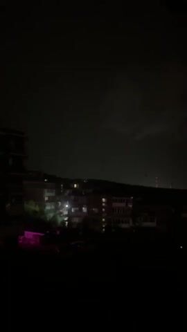 The video shows a nighttime view of buildings and a dark sky. There are lights on in some of the buildings. In the sky, there is a flash of lightning. The lightning appears to be distant and illuminates the clouds. The video is dark and grainy, making it difficult to see fine details.