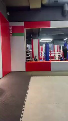 Here's a description of the video:
The video shows the interior of a boxing gym, with a clear view of a boxing ring. A person wearing boxing gloves and athletic gear is sitting inside the ring. The person is holding a phone and appears to be taking a selfie or photo of themselves. The boxing ring is red, white, and blue. The gym has a white and gray floor with red trim on the walls. The background is slightly blurred.