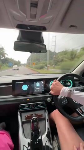 The video shows the interior of a car driving on a road. The dashboard display shows a circular graphic and a music interface with icons. The steering wheel is visible with a person's hands on it. Outside the car, a road with other vehicles and green hills with trees can be seen under a cloudy sky. A sign with Thai text is visible on the side of the road. The camera appears to be mounted inside the car, facing forward.