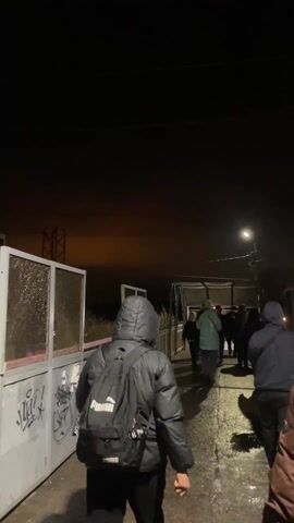 The video depicts a group of people walking along a covered walkway on a dark night. A person in a black hooded jacket and black pants, wearing a black Puma backpack, is in the foreground, walking away from the camera. Other people are ahead, some wearing jackets and hats, walking in the same direction. The walkway has a metal fence with graffiti on one side and a roofed structure overhead, illuminated by a streetlight. The ground is wet, with puddles visible. In the distant background, there's a partially visible metal structure and a dark sky with a faint orange glow on the horizon. The camera follows the person with the Puma backpack, maintaining a steady, slightly shaky viewpoint. The overall atmosphere is dark and somewhat bleak.