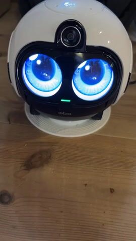 A close-up shot shows a white robot with large, bright blue digital eyes and a camera lens above them. The robot is sitting on a white circular base, which rests on a wooden surface. A green light is visible below the robot's eyes. The background is slightly out of focus, showing a dark object and a black cable. The robot appears to be stationary.