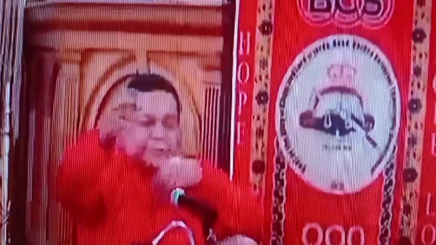 The video shows a man in a red shirt speaking into a microphone. He is seated behind a wooden podium. To his right is a large red banner with a circular emblem and the word "HOPE" printed vertically. The emblem appears to be a crown over an image with a sword and a motto. The background shows wooden paneling. The lighting is reddish and seems to be from stage lights. The man gestures with his right hand as he speaks. The overall scene appears to be a public address or performance.