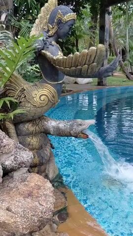 A fountain in the shape of a mythical creature pours water into a swimming pool. The creature has the upper body of a human with wings and the lower body of a bird. The water flows from the creature's mouth, which is shaped like a dragon or serpent's head. The fountain is surrounded by rocks and lush green foliage, creating a natural and tranquil atmosphere. The swimming pool is a vibrant blue, reflecting the surrounding greenery. The sun casts dappled light through the trees, adding to the serene ambiance.