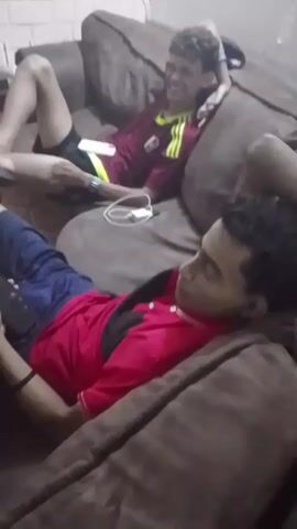 Two young men are relaxing on a brown sofa in what appears to be a home setting. The man in the foreground is lying down, dressed in a red polo shirt and blue shorts, looking down at a phone he holds in his hands. Behind him, another man is reclining with his head resting on his hand, wearing a maroon and yellow soccer jersey and dark shorts. He's looking towards the first man with a slight smile. A white electronic device with a cable is visible on the sofa between them. The wall behind the couch is painted white.