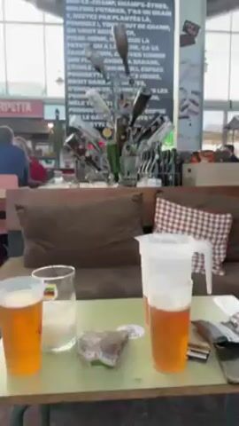 The video shows a brightly lit indoor market or food hall. Foreground: a table with drinks and food packaging, and a comfortable seating area with pillows. In the middle ground, an artistic bottle rack stands in front of a large chalkboard with text written in French. In the background, other stalls and patrons are visible.  The camera pans up to reveal the high, industrial-style ceiling featuring lighting fixtures and a wheelbarrow suspended as decoration.