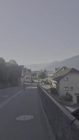 A vertical, long shot shows a street with houses and mountains in the background. The sky is overcast and hazy. A sidewalk runs along the right side of the street, separated from it by a low stone wall. A manhole cover is visible on the sidewalk. Trees and houses line the street.