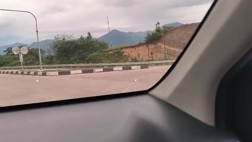 The video shows a view from inside a car driving on a highway. The road is bordered by a white guardrail with alternating black and white blocks. A hill with exposed soil is visible to the side. There are mountains in the background under a cloudy sky.  A road sign and another car are seen further down the highway. The view is through the driver’s side window.