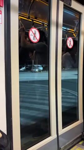 From a static camera position, the video depicts two doors on a bus or tram. Each door has a "No Leaning" sign prominently displayed. The doors are tinted, but it's possible to see figures standing inside. On the left, there appears to be a man holding onto a handrail. Another figure in a greenish shirt stands beside him. On the right side, an individual in a light-colored shirt can be seen. Outside the vehicle, the reflections suggest it is either nighttime or a dimly lit environment with striped lines on the ground.