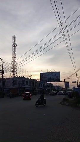 The video shows a street scene at dusk. A tall communication tower is visible to the left of the frame, standing beside several two-story buildings. Overhead, a network of power lines crisscrosses the sky. In the center of the view is a four-way intersection with cars and motorcycles moving through it. A large billboard promoting a university is positioned above the intersection. The sky is filled with a mix of light gray clouds and a bright orange glow from the setting sun, which is visible on the horizon. A few individuals can be seen on motorcycles, and some pedestrians are walking along the sidewalks. The environment suggests an urban or suburban setting in the evening light.