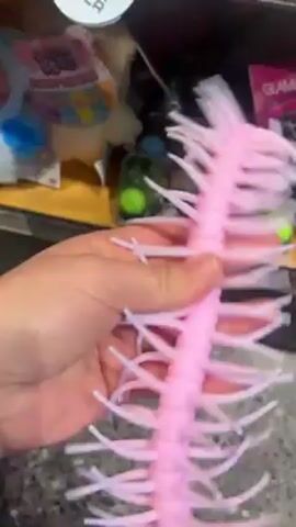 The video shows a person's hand holding and wiggling a pink and white toy. The toy has a segmented pink body with many white, spiky protrusions on each side, resembling a centipede or caterpillar. The hand holding the toy is visible against a background of a grey speckled surface and a shelf containing various other items, including a doll, a packaged toy, and some small balls. The hand manipulates the toy, showing its flexibility and texture.