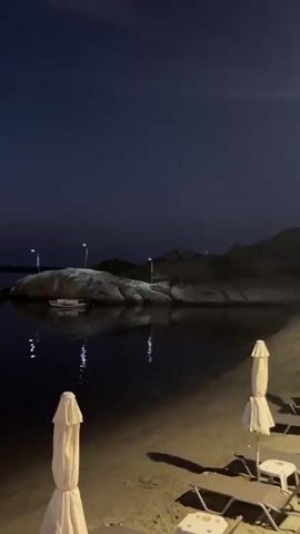 The video captures a twilight scene at a beach. The sky transitions from a dark blue to a lighter, orange-tinged hue near the horizon, where silhouetted mountains form a distant backdrop. The calm water reflects the lights from the shore and from what appear to be street lamps on a rocky outcrop in the middle ground. A small white boat is visible near the rocks. In the foreground, the sand of the beach is visible, along with the tops of two white beach umbrellas and a beach chair. The overall impression is one of a peaceful, dimly lit evening by the water.