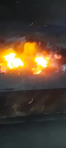 The video shows a close-up view of a fire burning with charcoal. The flames are bright orange and yellow, with some sparks visible. The charcoal pieces are dark and irregular in shape. The fire appears to be contained within a dark, possibly metal, enclosure. The overall impression is one of intense heat and combustion.