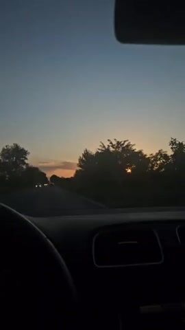 The video shows a driver's perspective from inside a car. The car is driving on a road, and a sunset is visible in the distance through the front windshield. The sky is a mix of blue and orange hues. Trees line the sides of the road, appearing as silhouettes against the bright sunset. Other cars are on the road ahead. The interior of the car is visible, including the steering wheel and dashboard. Music is playing in the background.