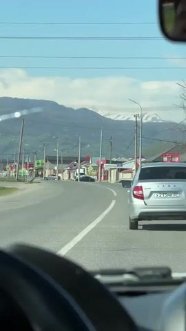 The video shows a view through the windshield of a car moving on a road, with a silver Lada car in front. The Lada's license plate number is X215MK 07. The road is surrounded by buildings, utility poles, and mountains in the distance. There's a sign indicating "ЗАЮКОВО" (Zayukovo) visible on the right side of the road. The sky is blue with some clouds. The camera captures a partial reflection of a person's face in the rearview mirror. The car is traveling along the road on a sunny day.