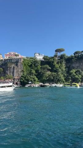 A video taken from a boat shows a body of water with a cliffside town in the background. The town is built on a steep cliff and features a mix of colorful buildings, some with balconies and arches. Several boats are docked near the cliff, including a white passenger boat in the foreground and smaller boats and watercraft further down the coast. The water is a vibrant turquoise, and the sky is a clear, bright blue. Trees and lush greenery cover the cliffside, creating a natural contrast with the man-made structures. The overall scene depicts a scenic coastal town on a sunny day.