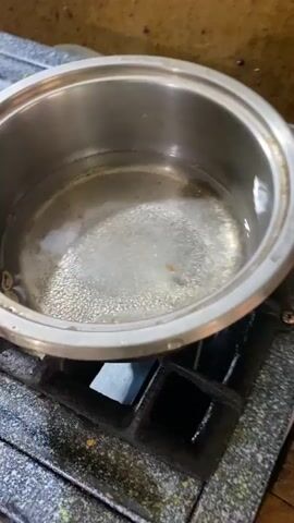A metal pot of water is seen boiling on a gas stove. The water is bubbling vigorously, and a small, dark object is floating in the center of the pot. The stove appears to be old and worn, with some rust and grime visible. The background is dimly lit and out of focus, but it appears to be a kitchen environment. A wooden cutting board and a pink object, possibly a utensil, are visible on the right side of the frame.