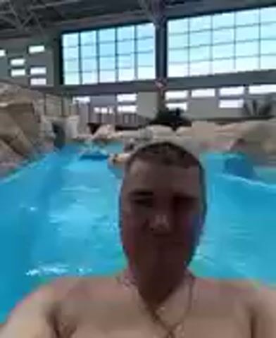 A man in his 30s with short dark hair and a beard is submerged in a bright blue body of water, possibly a wave pool or lazy river. He is wearing a necklace. The water appears to be indoors, as evidenced by the ceiling structure above him, which features large rectangular windows or skylights and industrial beams. To his left and in the background, there are artificial rock formations with a textured, layered appearance. The water is calm in the immediate foreground, with small ripples visible. The man's chest and shoulders are visible, and he is looking directly at the camera with a slight smile. The perspective suggests the video is a selfie taken from within the water.