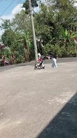 The video shows two young girls on a paved area next to a road. The girl on the left is wearing a white hijab and dress and is standing next to a baby stroller containing a child. The girl on the right is wearing a dark hijab and a white and blue patterned outfit and is pushing the stroller. The background shows trees, houses, and a power pole. Indonesian flags are visible on poles along the road. A motorcycle is seen in the background on the road. The weather appears to be sunny.