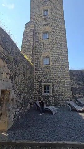 This video captures a tour of Stolpen Castle in Germany. The scene opens with a view of a tall, stone tower, likely part of the castle's fortifications. A young person in a black t-shirt is visible in the foreground, walking across a gravel area. In the background, there are several curved wooden benches. The weather appears to be clear and sunny, suggesting daytime.

The camera then pans to show a section of the castle wall made of rough, grey stones, with some vegetation growing along the top. An arched stone passageway is also visible. The perspective shifts to look through this archway, revealing a wider view of the castle grounds. Another stone tower, smaller than the first, stands in the distance with a green flag flying from its top. A wall encircles the courtyard, and the ground is covered in gravel and a paved path. There are a few other people visible in the distance. The overall impression is of exploring a historical site on a pleasant day.