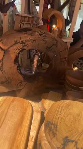 The video pans across a display of wooden handcrafted items, likely at an outdoor market or craft fair. In the foreground, several wooden cutting boards with handles are stacked. Their surfaces feature natural wood grain and intricate dark patterns that resemble lightning strikes, possibly from a technique like Lichtenberg figure burning. To the left, a large wooden bowl and a carved wooden lamp with exposed bulbs are visible. In the background, a person, likely a vendor, is partially visible behind a table laden with more wooden bowls and other crafts. They appear to be interacting with the items on the table, and possibly a customer. The environment suggests an outdoor setting with natural light and a tent or canopy structure providing shade. A pink and white patterned ball rests on one of the wooden surfaces.