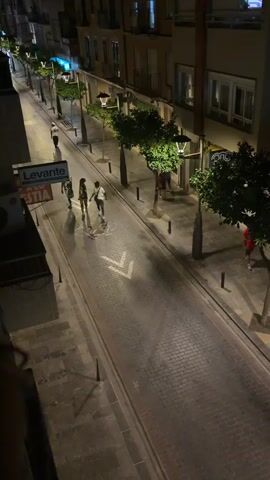 A nighttime, elevated view shows a cobblestone street in a European-style urban environment. The street is lined with buildings, trees, and streetlights, some of which are illuminated. A group of four young men are in the middle of the street, walking from left to right in the frame. Two of the men are wearing dark clothing, one is wearing a white shirt, and another is wearing a patterned shirt. They appear to be interacting and walking together. In the background, a few other figures can be seen on the sidewalk, and some storefronts are visible. The lighting suggests it is after dark.