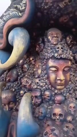 A close-up video shows a sculpture made of numerous human faces and skulls. A central, golden-colored face with closed eyes is prominent. Surrounding this face are many smaller faces and skulls, rendered in dark, metallic colors. To the left and right of the main sculpture are large, blue, horn-like or tusk-like objects. The sculpture appears to be part of a larger, dark structure. The lighting is dim, highlighting the metallic sheen of the sculpture.