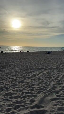 The video shows a beach scene during sunset. The sun is low in the sky, casting a warm glow and its reflection on the ocean. The foreground is dominated by sand dunes and ripples. In the middle ground, several people are scattered along the shoreline, some sitting in beach chairs, others standing or walking. A few beach chairs and umbrellas are visible. The ocean is calm with gentle waves. The sky is partly cloudy with streaks of light. The camera pans slightly to the left and right, capturing different groups of people and their activities, like someone walking near the water's edge and a group of people gathered with chairs. The overall atmosphere is peaceful and relaxed.