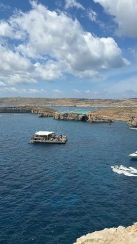 A wide-angle shot taken from a high vantage point shows a sunny coastal scene. The sky is blue with scattered white clouds. Below, the deep blue sea stretches out to the horizon, marked by a rugged coastline in the distance. A floating structure with a white roof sits in the middle of the water, and a speedboat is cutting through the waves, leaving a white wake behind it. The foreground of the shot includes a rough, rocky outcrop.