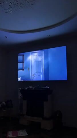 A man in a dark robe opens a door and enters a room. Another man in a dark suit with a purple sash enters the room. They stand facing each other. The scene is projected onto a large screen mounted above a fireplace. The room is dimly lit, with a patterned wallpaper and a curved ceiling.