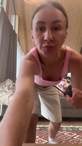 A woman in a pink crop top and white shorts dances in a room with wood-paneled walls and a pitched ceiling. She is in the foreground, with a couch and luggage in the background. She jumps, raises her arms, and makes excited gestures. Another person is partially visible in the background.