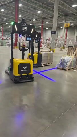 The video shows two yellow autonomous pallet jacks in a large warehouse. The jack in the foreground has the number 107 on it.  The jacks each shine two parallel blue laser lines, one on each side, to the front.  There are pallets loaded with goods in the background of the warehouse, and the floors are polished cement.  The jack in the front has the name "ABTOMAKOH" printed on the front of it.