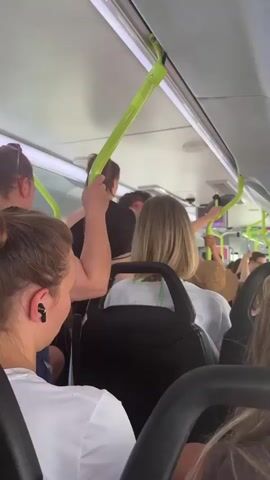 The video begins inside a crowded bus, showing passengers holding onto bright green poles for support. The camera then shifts to a view through the bus window, revealing a street scene. Several bicycles are parked on a sidewalk next to a tree. A yellow sign with a black outline and the number "5" inside is mounted on a pole. A white truck with red and white reflective tape on its rear is partially visible. A person on a bicycle is seen riding away from the truck. The buildings lining the street appear to be residential, with brick facades and windows. The sun is shining, casting shadows.