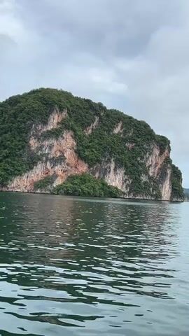 A drone shot surveys a body of water with a large, lush, green island with rocky cliffs in the foreground, gradually moving away from the island to reveal more islands in the distance under a cloudy sky.