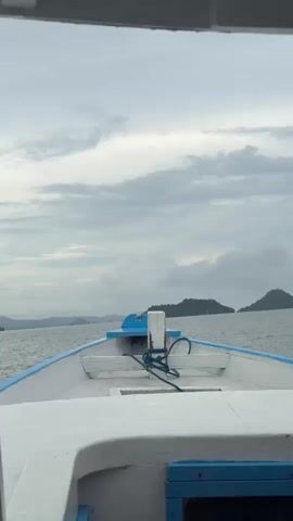 The video shows the front view of a small white and blue boat sailing on a body of water. The sky is overcast with gray clouds, and in the distance, there are islands or landmasses visible. The boat is moving forward, and the water is slightly choppy. There are no people visible in the frame. The focus is primarily on the boat's movement through the water and the scenic view of the landscape. The video appears to be taken from inside a larger boat or structure, as indicated by the frame at the top of the shot.