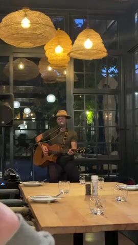 A man with a hat is playing guitar and singing into a microphone. He is sitting between a long table that is set for a meal, and a large window. There is a speaker to his left.  Above the table are several large, round lamps made of woven material. Beyond the window are trees, lights, and the dimming sky.