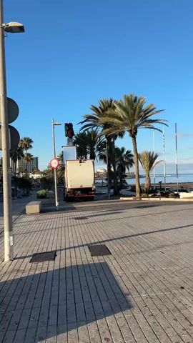 This media file captures a scene in Adeje, Spain, likely in the early morning given the low angle of the sun and the long shadows cast across the paved pedestrian area. The sky is a clear, bright blue, indicating fair weather.

The primary subject is a white truck with a beige rear compartment, possibly a waste disposal or service vehicle, parked on the street. Mounted on top of the truck is equipment that includes what appears to be a traffic light and a camera system. A circular red and white sign with a bicycle symbol is visible on a pole nearby, suggesting a designated cycling path or a restriction for bicycles.

The setting is a coastal promenade. Palm trees are scattered throughout the scene, with some larger, mature ones framing the right side of the frame. In the background, the calm sea is visible, and further in the distance, buildings line the coast, suggesting a town or resort area. The pedestrian area is paved with light-colored rectangular tiles laid in a patterned fashion, with metal grates embedded in the surface. A tall street lamp stands to the left of the frame.

The overall impression is one of a quiet, functional operation taking place in a scenic, sunny location, likely as the day begins. There are no people immediately visible in the frame. No discernible text is visible on the truck or any other prominent objects.