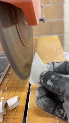 A close-up shot shows a gloved hand holding a white tile fragment against a spinning circular saw blade. The saw is orange and gray and appears to be a wet saw, as there is water visible on the surface. The tile fragment is being cut into a smaller piece. Sawdust and small tile fragments are scattered on the orange surface of the saw. The background is out of focus, but it appears to be a brick wall.