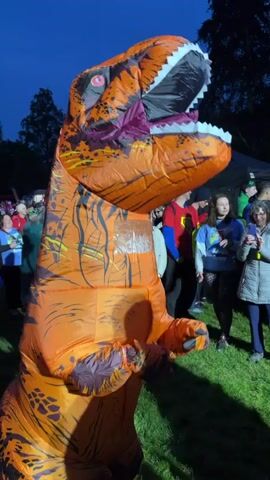 The video shows a person in an orange T-Rex inflatable costume standing in a crowd of people. The T-Rex is patterned with dark lines. People are standing around, some are taking photos with their phones. There is a white tent and trees in the background. It is dusk or night, and it appears to be an outdoor event.