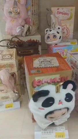 The video shows a close-up view of a display case filled with various toys and collectibles. The items are predominantly plush toys and figurines with large, expressive eyes and often feature anime-inspired designs. Many of the items are packaged in colorful boxes with artistic illustrations. There are keychains, dolls, and larger plush creatures visible. The lighting suggests an indoor retail environment, possibly a toy store or a section within a department store. The camera pans and zooms slightly, providing a detailed look at the merchandise. Some of the packaging has text and prices displayed, indicating that these are items for sale. The focus remains on the toys, with no people directly visible in the frame.