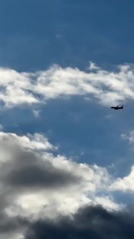 This is a vertical, medium shot video of a commercial airplane silhouetted against a bright blue sky filled with dramatic, puffy white and gray clouds. The airplane is positioned in the upper right quadrant of the frame, traveling from left to right. The clouds are varied in texture and density, with some appearing wispy and others heavy and shadowed, suggesting a dynamic weather system.

The scene is an outdoor aerial view, likely captured from the ground looking upwards. The lighting and cloud formations suggest it is daytime, possibly late afternoon or early morning given the contrast between the bright sky and the shadowed clouds, and the potential for dramatic lighting. There are no people or specific objects visible besides the airplane.

The location is identified as Pyachanki, Russia. This detail, combined with the image of a commercial aircraft, implies the airplane is in transit over or near this Russian location. The overall mood is one of vastness and movement, with the airplane a small but significant element within the expansive sky. There is no visible text in the media file.