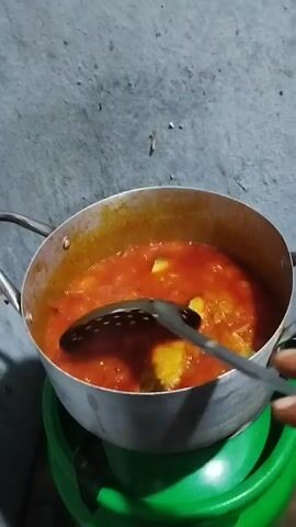 A person stirs a pot of bright red sauce with large pieces of light yellow vegetable, likely squash, in it. The pot is metal, likely aluminum, and sits on top of a green plastic stool. The background is a plain concrete wall. The person uses a slotted spoon with a grey handle. The sauce simmers and splashes with each stirring motion. The scene is close up and well lit.